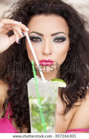 Portrait of pretty young brunette woman with curly hair and bright makeup drinking alcoholic mojito cocktail from mint soda light rum ice cubes and lime with straw, vertical picture