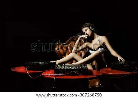 Attractive young glamour dj woman in lingerie and headphones sitting at red table with mixer console on brown leather royal chair in night club on dark background, horizontal picture