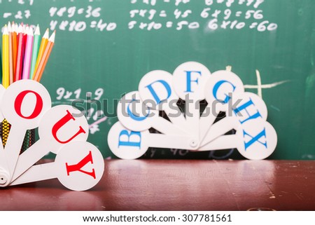 Colorful pencils of red yellow orange violet purple pink green and blue in stationary cup ruler and fan english alphabet standing on brown school desk on written with white chalk blackboard on math