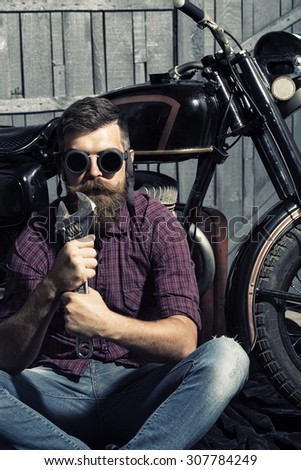 Young unshaven man biker in purple checkered shirt and glasses sitting near motorcycle in garage holding metallized iron wrench looking forward on workshop background, vertical picture