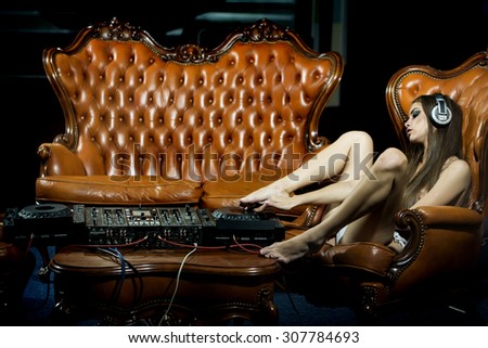 Sensual sexy undressed dj girl in headphones with bare chest sitting on chair at table with mixer console and brown leather royal sofa in night club copyspace, horizontal picture