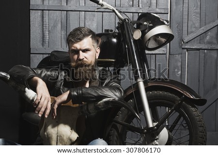Thoughtful unshaven male biker in leather jacket sitting near motorcycle in garage with big bone skull antlers of stuffed animal looking forward on wooden wall background, horizontal picture