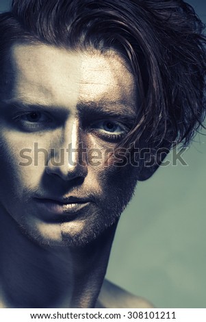 Closeup of young fashionable painted man model with gold bodyart on one half of face and stylish hairdo looking forward standing in studio on grey background, vertical picture