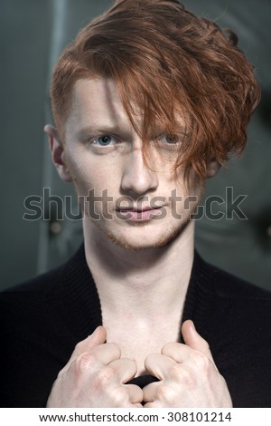 Portrait of attractive stylish young unshaven guy model with red hair holding collar with hands standing in black jersey looking away indoor on studio background closeup, vertical picture