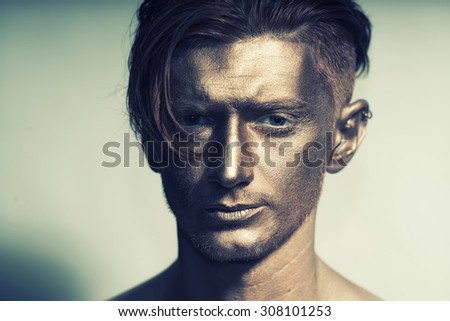 Closeup of young fashionable painted man model with bronze bodyart on face and stylish hairdo looking forward standing in studio on white background, horizontal picture