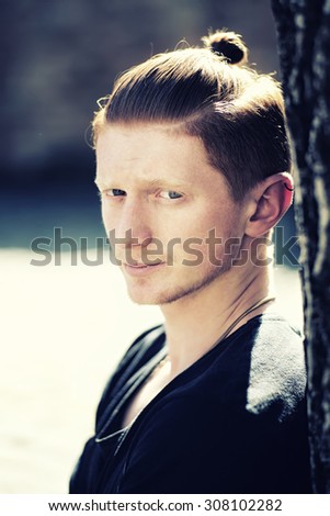 Portrait of young sexy stylish handsome unshaven man with red hair in ponytail in black clothes with small ear ring standing near tree outdoor sunny day, vertical picture