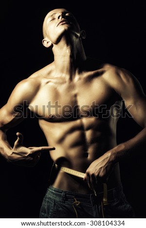 Closeup of young naked male in jeans with sexual strong muscular beautiful tan body showing finger on yellow tape measure round waist standing on black background, vertical picture