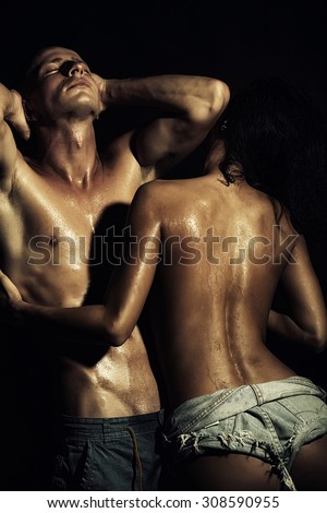 Sexy young pair of naked man with strong muscular beautiful wet body and pretty slim tan girl in jeans shorts embracing standing on black studio background, vertical picture