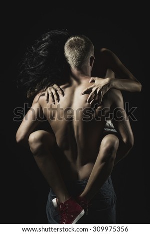 Young naked couple of sexy woman in red trainers sitting on man with strong muscular body standing with back on black studio background, vertical picture