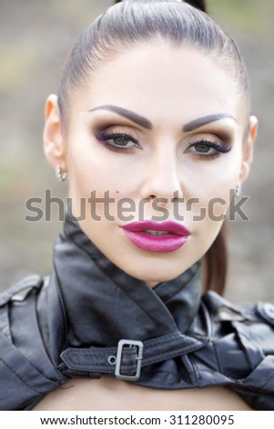 Portrait of cool beautiful brunette young serious woman with bright make-up and ponytail in black leather jacket looking forward outdoor on natural background, vertical picture