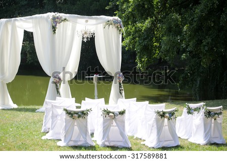 Beautiful decorated with light white chiffon chandelier and bouquets of roses pastel colors wedding pavilion chairs and table standing on green grass near lake and trees sunny day, horizontal picture