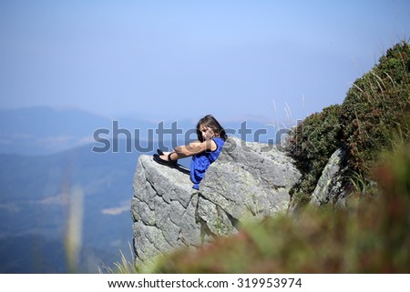 Pretty brunette thoughtful girl with long hair in blue lace dress sitting on stone rock cliff in beautiful landscape sunny day outdoor on natural blue sky background, horizontal photo