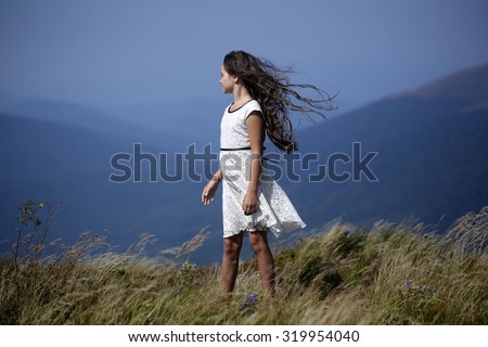 Little pretty brunette girl in white lace summer dress looking away standing in mountain valley with deep dry spikelet grass sunny windy day outdoor on natural blue background, horizontal picture