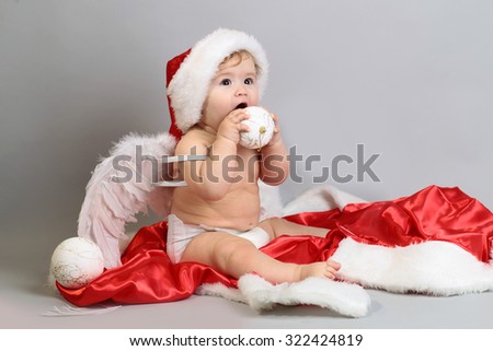 One small beautiful blonde boy child with curly hair in angel wings with feathers red hat and coat of santa playing with traditional decorating new year tree balls sitting in studio on grey background