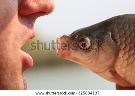Closeup view of human male face and fresh fish head with open mouth looking at each other in funny dialogue sunny day outdoor on natural background, horizontal picture