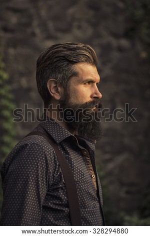 Closeup side view of one handsome senior man with black hair and long lush beard in blue shirt and purple trausers standing outdoor on stone wall grey background, vertical picture