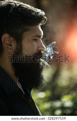 Closeup portrait side view of one senior serious man with long lush beard holding in mouth and smoking unhealthy cigarette with smoke sunny day outdoor on natural background, vertical picture