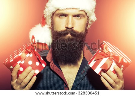 Portrait of one senior new year man with long black beard in shirt and red santa claus hat holding two wrapped present boxes in hands for christmas standing in studio on purple background, horizontal