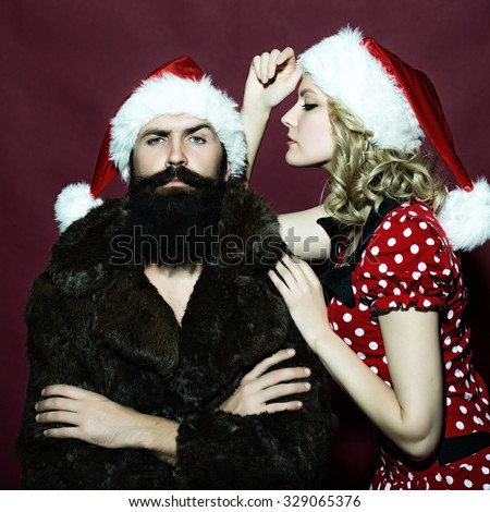 New year young couple of blond woman with curly hair in dress and man with long beard in red santa claus hat and fur coat celebrating christmas standing on studio purple background, square photo