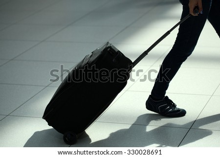 Closeup back view of female legs in jeans with black rolling suitcase wheeled travel bag trolley case moving over airport terminal floor tile background, horizontal picture