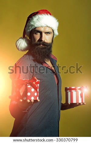 One handsome senior new year man with long black beard in shirt and red santa claus hat holding two wrapped present boxes in hands for christmas standing in studio on yellow background, vertical photo