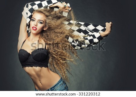 Portrait of attractive radiant looking blond young woman with fly-away wavy long hair wearing black and white cardigan underwear bra jeans posing in studio on grey background, horizontal picture