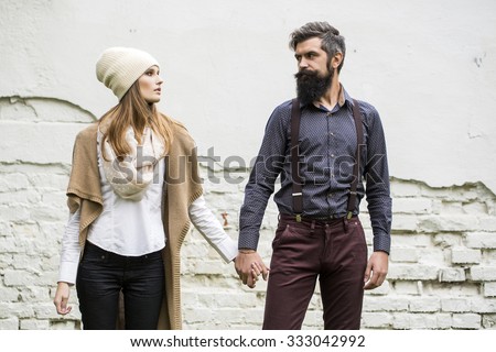 One beautiful stylish emotional couple of young woman and senior man with long black beard standing close to each other outdoor in autumn street on white brick wall background, horizontal picture