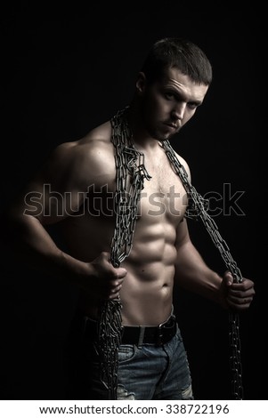 One handsome sexual strong young man with muscular body in blue jeans holding chain with hands hanging on neck and shoulders standing posing in studio on black background, vertical picture