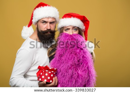 Young beautiful happy new year couple of man with long beard with present box and woman in red santa christmas hat with fur in studio on yellow background, horizontal picture