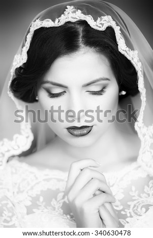 Portrait with side view of one beautiful young brunette sensual pensive bride in lace dress and veil on head holding hands near face black and white, vertical picture