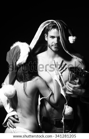 Young sexual new year couple of woman in corset and christmas hat embracing muscular man in santa claus winter coat holding wine bottle with cork in pail in studio black and white, vertical