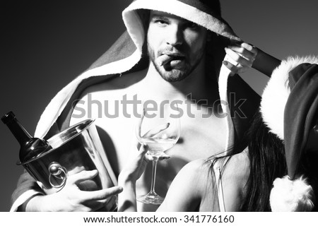 Young sexual new year couple of woman in corset and christmas hat embracing muscular man in santa claus winter coat holding wine bottle with cork in pail in studio black and white, horizontal