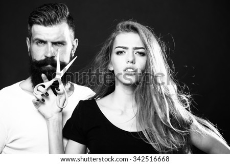 Closeup view of young beautiful couple of beautiful fashionable female barber cutting and holding scissors and handsome man with long beard in studio black and white, horizontal picture