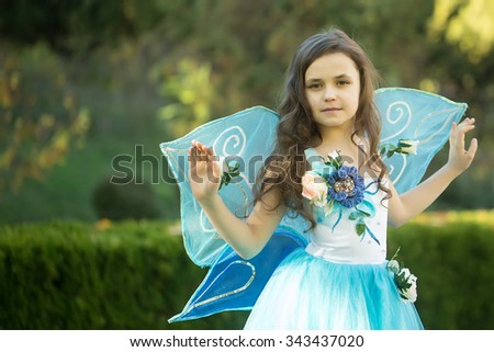Pretty white small girl with loose curly dark hair in blue fluffy dress decorated with flowers and fairy wings posing at afternoon in park, horizontal photo