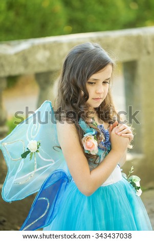 Closeup photo of charming white little girl with curly loose dark hair dressed as fairies with wings keep hands folded in day outdoor, vertical picture