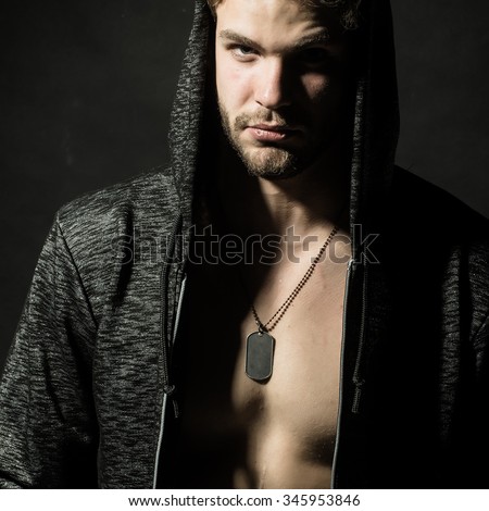 Portrait of one handsome confident serious young sexy man with strong body and muscular bare chest in jacket with hood and neck chain looking forward in studio on black background, square picture