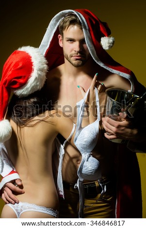 Young sexual new year couple of woman in corset and red christmas hat embracing muscular man in santa claus winter coat holding wine bottle with cork in pail in studio on yellow background, vertical