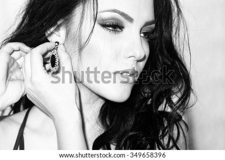Portrait of one beautiful young sexual woman with curly hair slim straight body and bright makeup in elegant dress putting on jewellery on ear standing indoor black and white, horizontal picture