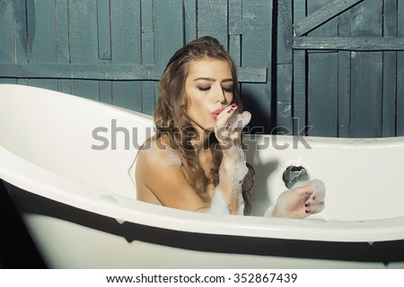 One beautiful sensual playful flirtatious young woman with long hair in blue knitted cloth sitting in white bath tub playing with soap foam indoor on wooden background, horizontal picture