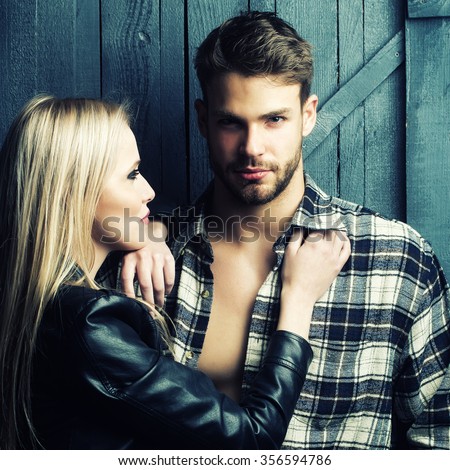 Young sexual couple of pretty blonde woman in leather jacket standing near handsome serious man in checkered shirt with bare muscular chest in studio on wooden background, square picture