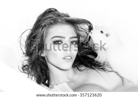 Portrait of one beautiful sensual pensive dreaming young woman with wet hair and bright makeup lying in bath tub with water and white soap foam looking forward black and white, horizontal picture