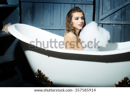 One beautiful sensual playful flirtatious young woman with long hair in blue knitted cloth sitting in white bath tub playing with soap foam indoor on wooden background, horizontal picture