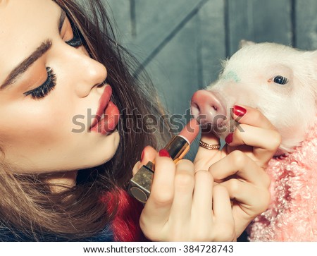 Beautiful young sensual fashionable woman holding cute pink small pig pet in cloth and lipstick in hands on wooden background, horizontal picture
