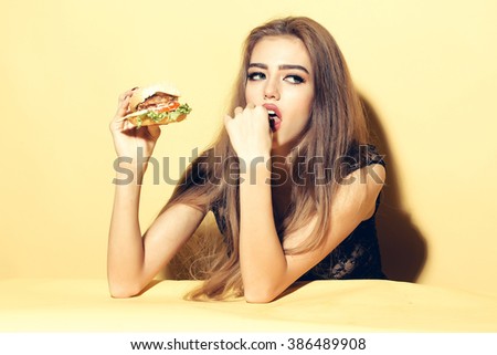 Beautiful young sensual woman eating tasty big fresh burger indoor on studio yellow background, horizontal picture