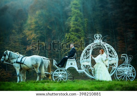 Young wedding romantic couple of bride in white dress and bridegroom in suit in cinderella carriage with horses in deep green forest outdoor on natural background, horizontal picture