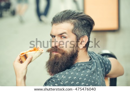 handsome sexy bearded young man hipster with long beard and mustache on serious hairy face eating pizza sitting on street bench outdoor