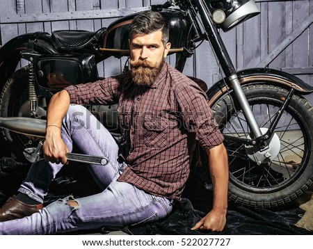 Bearded man hipster biker brutal male with beard and moustache sits on floor near motorcycle with wrench on wooden background