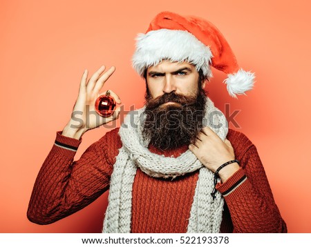 young handsome bearded santa claus man with long beard in red sweater and new year hat in scarf holds decorative christmas or xmas ball on orange studio wall background