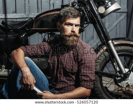 Bearded man hipster biker brutal male with beard and moustache in leather jacket sits on floor near motorcycle with wrench on wooden background