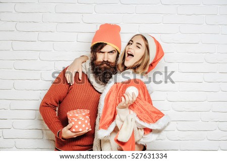 Young happy Christmas or new year couple of bearded man with gift box and pretty sexy girl or beautiful woman in red santa claus costume with xmas bauble and sock on white brick wall